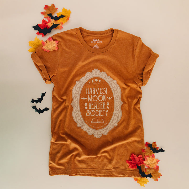 An orange-brown short-sleeve graphic T-shirt laid flat on a light beige background, surrounded by autumn leaves and small black bat cutouts. The shirt has a large cream decorative oval graphic on the front reading “Harvest Moon Reader Society.