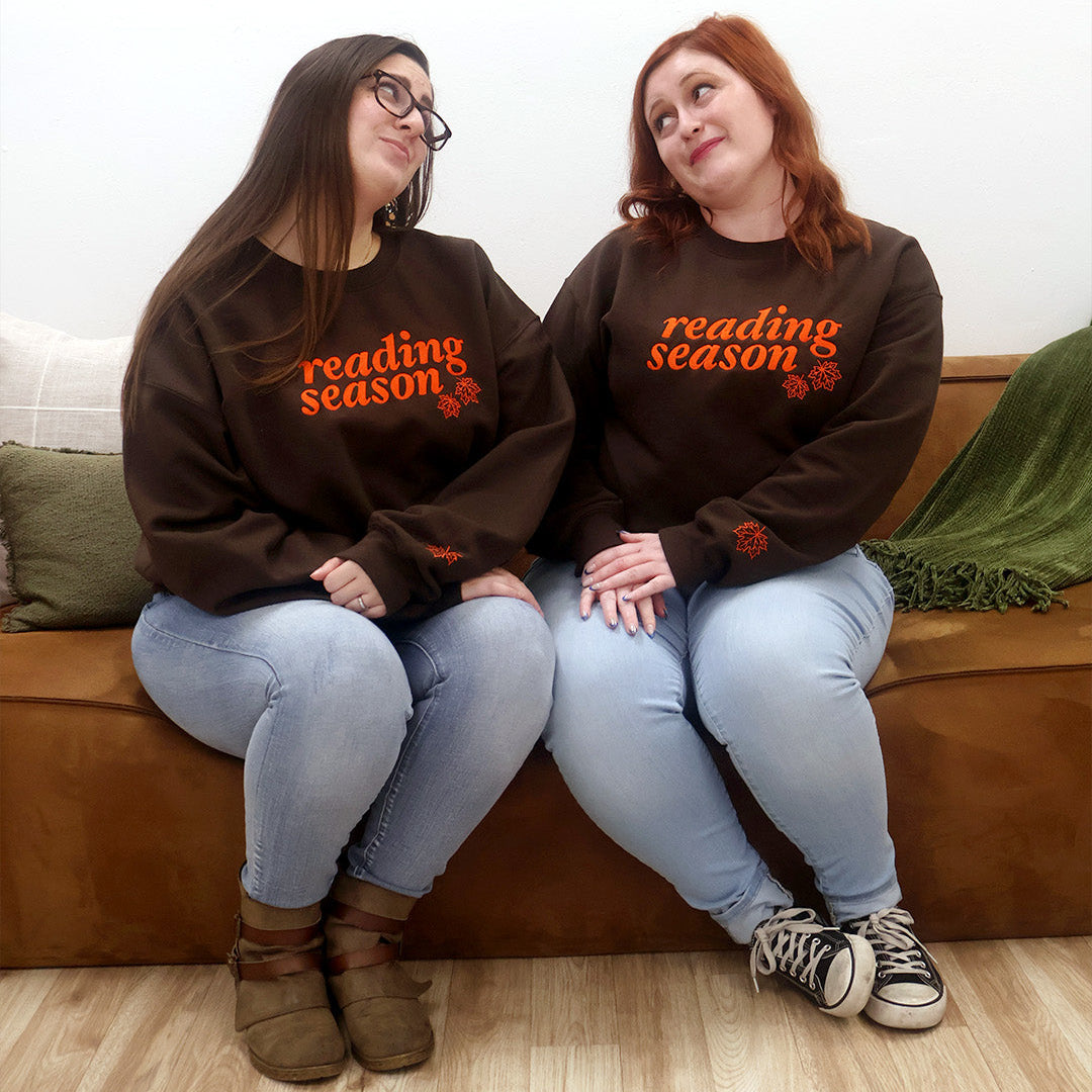 Two women sitting side-by-side on a brown leather couch, wearing matching dark brown crewneck sweatshirts with orange text that reads 'reading season' and a small orange leaf graphic.
