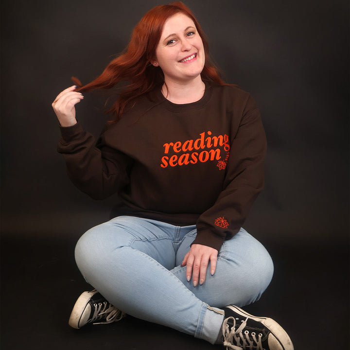 A smiling woman with red hair sits cross-legged on a black studio background, wearing a dark brown sweatshirt with orange text that reads 'reading season' and light blue jeans.