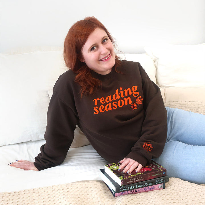 A smiling woman with red hair sits on a bed wearing a dark brown sweatshirt with 'reading season' printed in orange across the chest, paired with light blue jeans. She is holding a small stack of books.