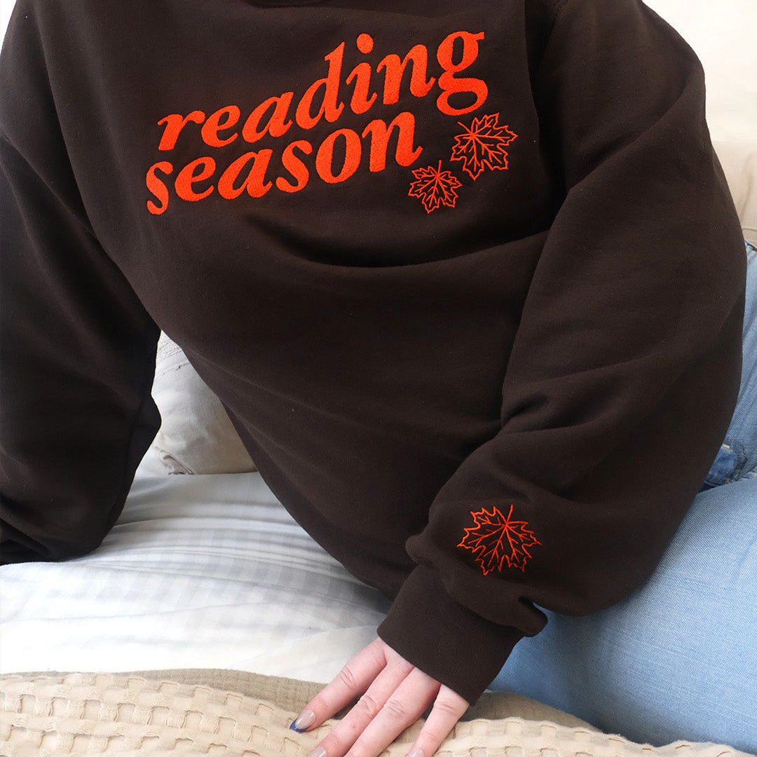 A woman wearing a dark brown crewneck sweatshirt with 'reading season' and leaf details printed in orange across the chest, paired with light blue jeans, sitting on a bed with a textured beige blanket.