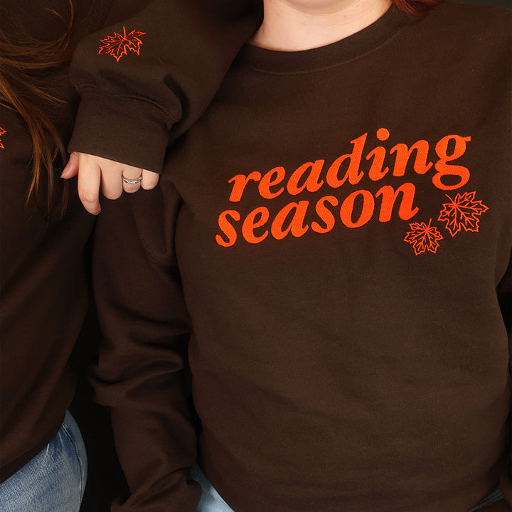 A woman wearing a dark brown crewneck sweatshirt with 'reading season' printed in orange across the chest, paired with light blue jeans. Another woman is partially visible on the left, also wearing a matching sweatshirt.
