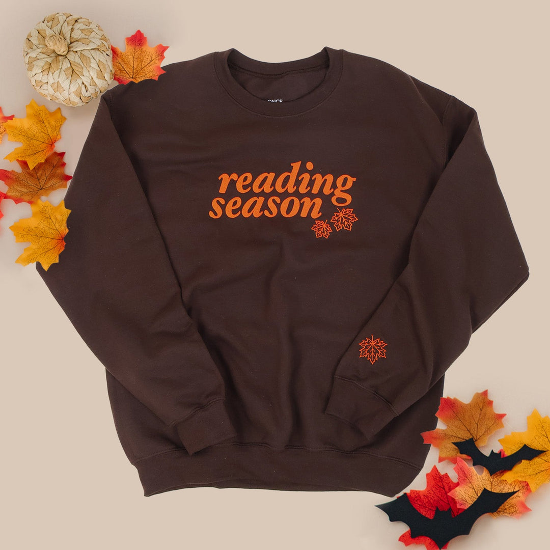 A dark brown crewneck sweatshirt with 'Reading Season' printed in orange across the chest, styled in a flat lay with autumn leaves, a small pumpkin, and bats on a beige background.