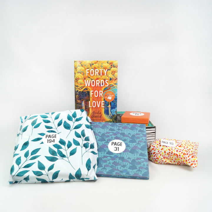 a hardcover edition of Forty Words for Love and an orange box sit on a green box. In front of the box are a white polybag with green leaves on it, a box with a pattern of houses, and a drawstring bag with a white/orange/red dotted pattern. The boxes and bags all have page numbers.