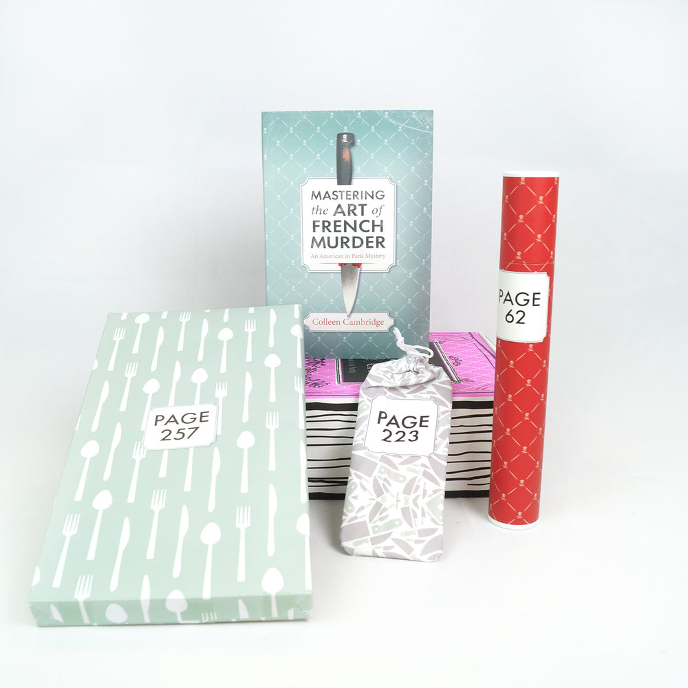 A hardcover special edition of Mastering the Art of French Murder stands on a pink box. In front are a rectangular box, small drawstring bag, and red tube. The boxes, tube, and bag all have page numbers.