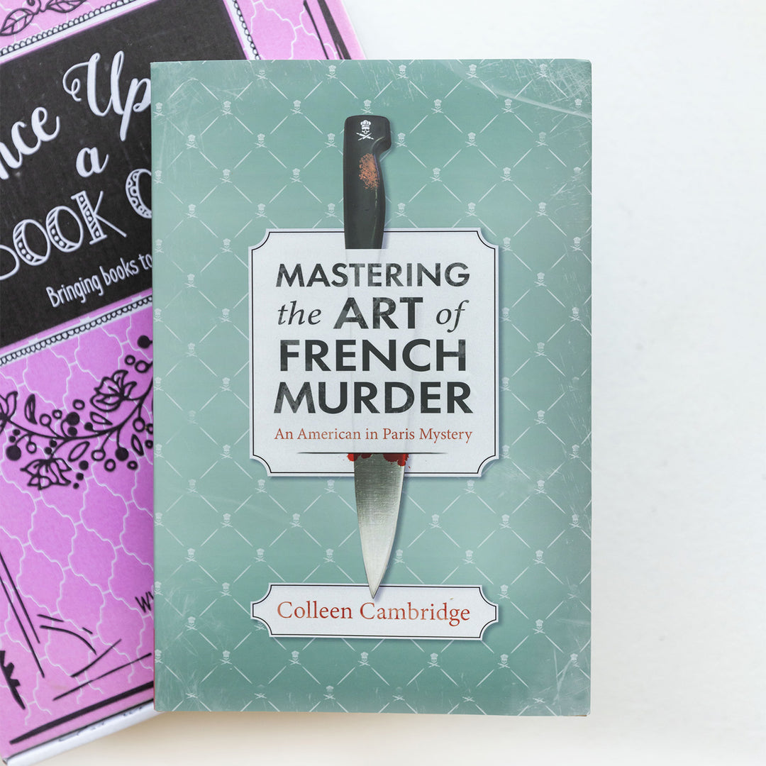 the front of a hardcover special edition of Mastering the Art of French Murder on top of a pink box. Cover shows a teal background with crosshatch pattern and a kitchen knife in the center