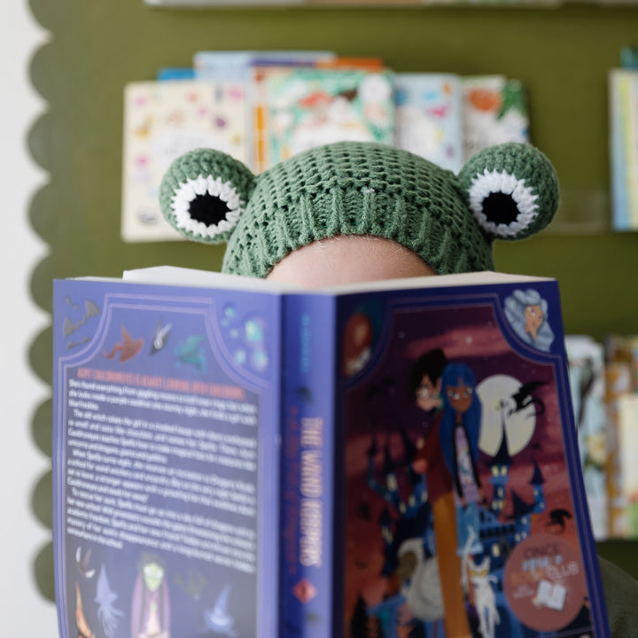 A child holding an open book in front of them wears a dark green crochet frog hat.