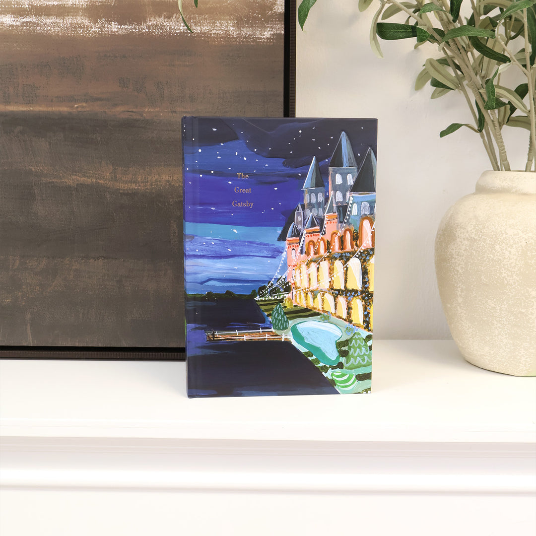 A full, deluxe painted edition of The Great Gatsby by F. Scott Fitzgerald featuring Laci Fowler's distinctive hand-painted artwork, enhanced with embossing and gold foiling, decorative interior pages with memorable quotes sprinkled throughout, and a matching ribbon marker and gold page edges for a sophisticated finish