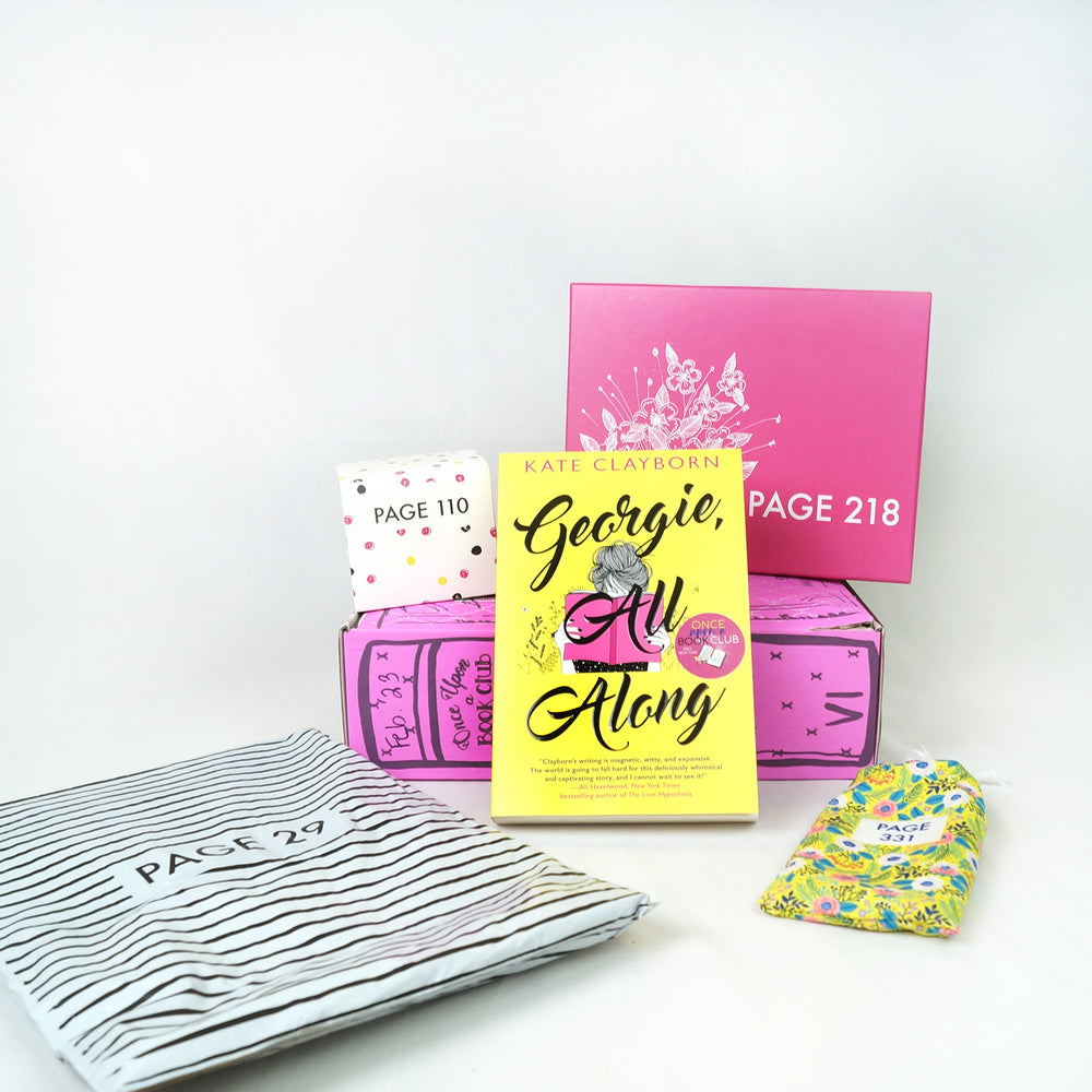 A paperback edition of Georgie, All Along leans against a pink Once Upon a Book Club box. On top of the pink box are a white square box and a pink box. In front of the book are a white and black striped bag and yellow drawstring bag. The boxes and bags all have page numbers.