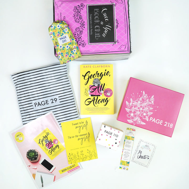 A pink Once Upon a Book Club box is at the top of the image with a yellow drawstring bag on it. In front of the box are a black and white striped bag, paperback edition of Georgie, All Along, a pink box, bookclub kit, quote card, white square box, bookmark, and signature card. The boxes and bags all have page numbers.