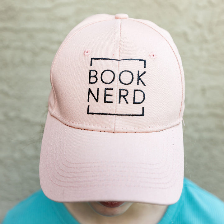 a white woman wears a pale pink baseball cap that says BOOK NERD in black writing on the front