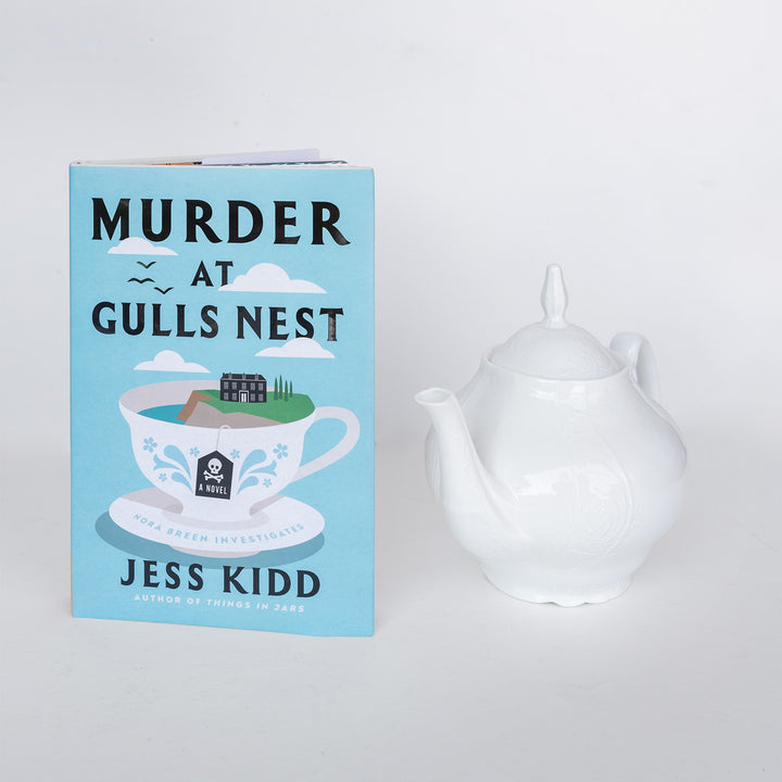 Book titled 'Murder at Gulls Nest' by Jess Kidd next to a white teapot on a light gray background