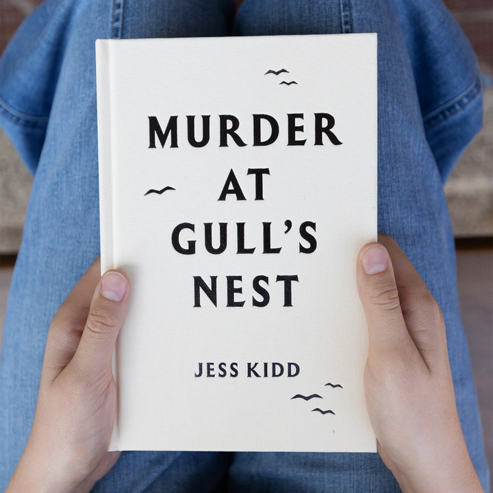 Book titled 'Murder at Gull's Nest' by Jess Kidd without it's dust jacket, held by a person wearing blue jeans.
