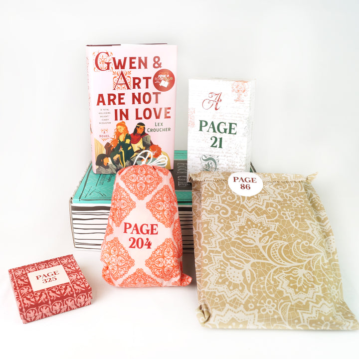 hardcover book "Gwen & Art Are Not in Love" by Lex Croucher and a white box with script on it sit on top of a green box. In front of the green box are a square red box, a red and pink drawstring bag, and a brown paper bag with white lace. The boxes and bags all have page numbers.