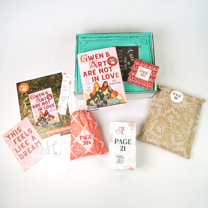 a hardcover copy of "Gwen and Art Are Not in Love" sits on a green box next to a square red box. In front are paper items (quote card, bookclub kit, bookmark, and signature card), a red and pink drawstring bag, a white rectangular box, and a brown rectangular bag with white lace. The boxes and bags all have page numbers.