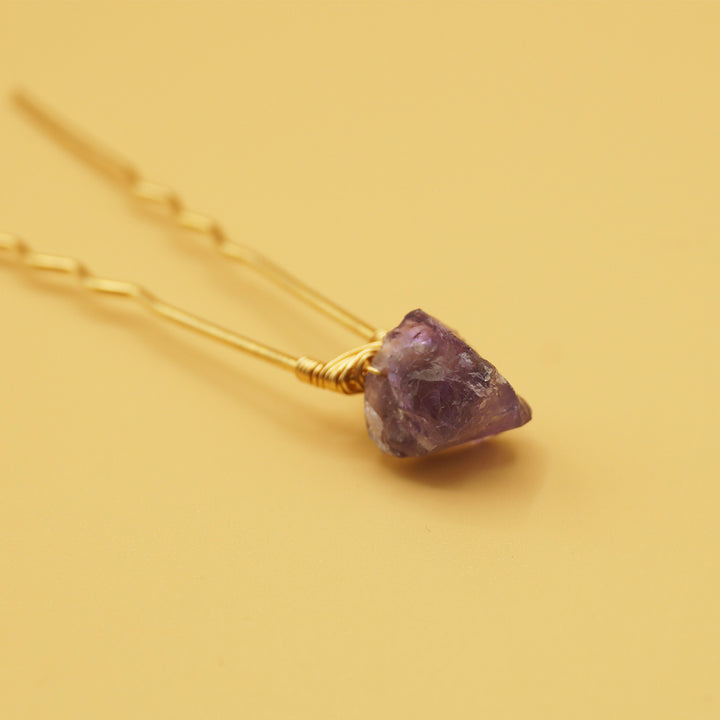 a hair pin lays open with a purple amethyst gemstone on it, against a yellow background