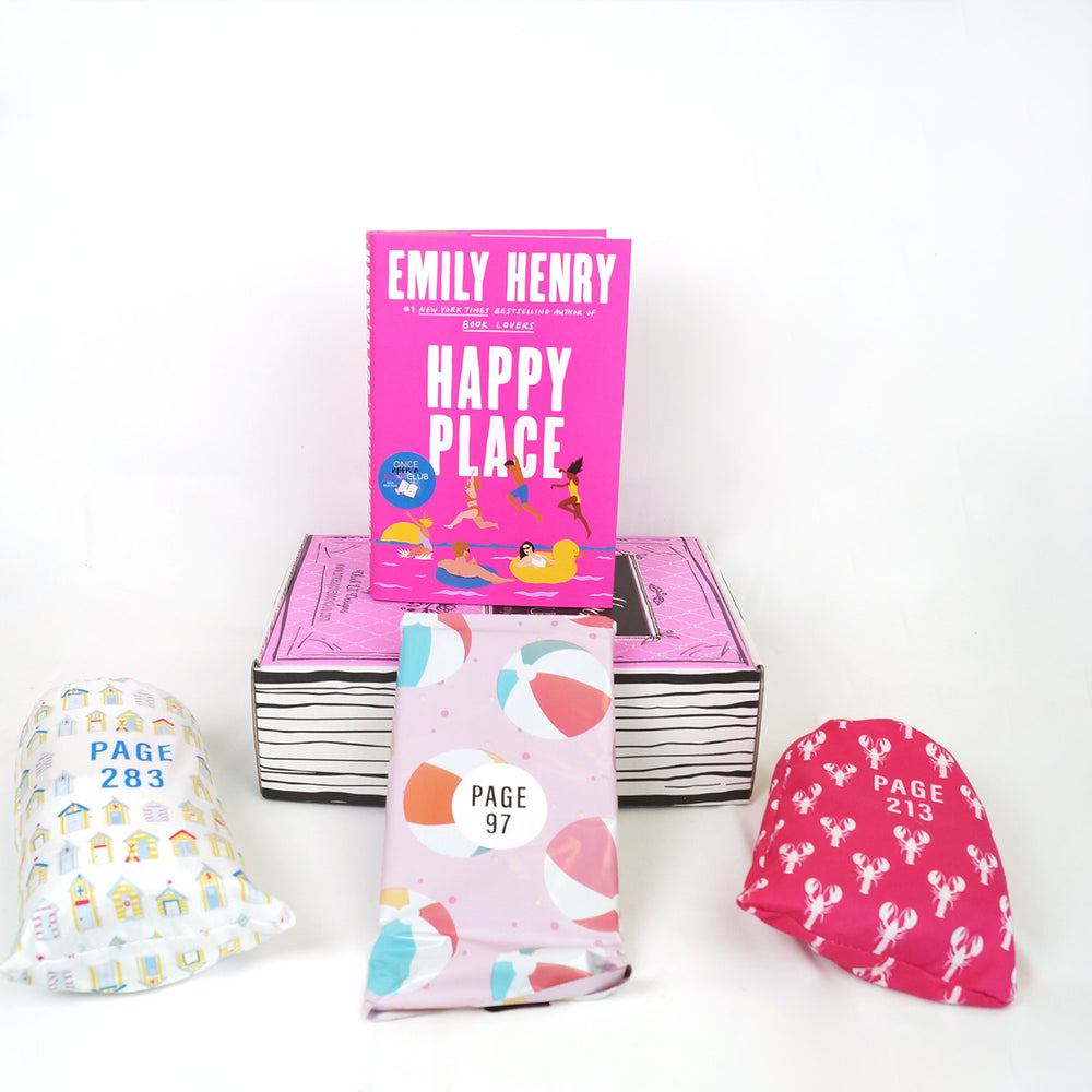 A hardcover edition of Happy Place sits on a pink Once Upon a Book Club box. In front of the box are a white drawstring bag with a pattern of houses, a pink polybag with beachballs on it, and a pink drawstring bag with lobsters on it. The boxes and bags all have page numbers.