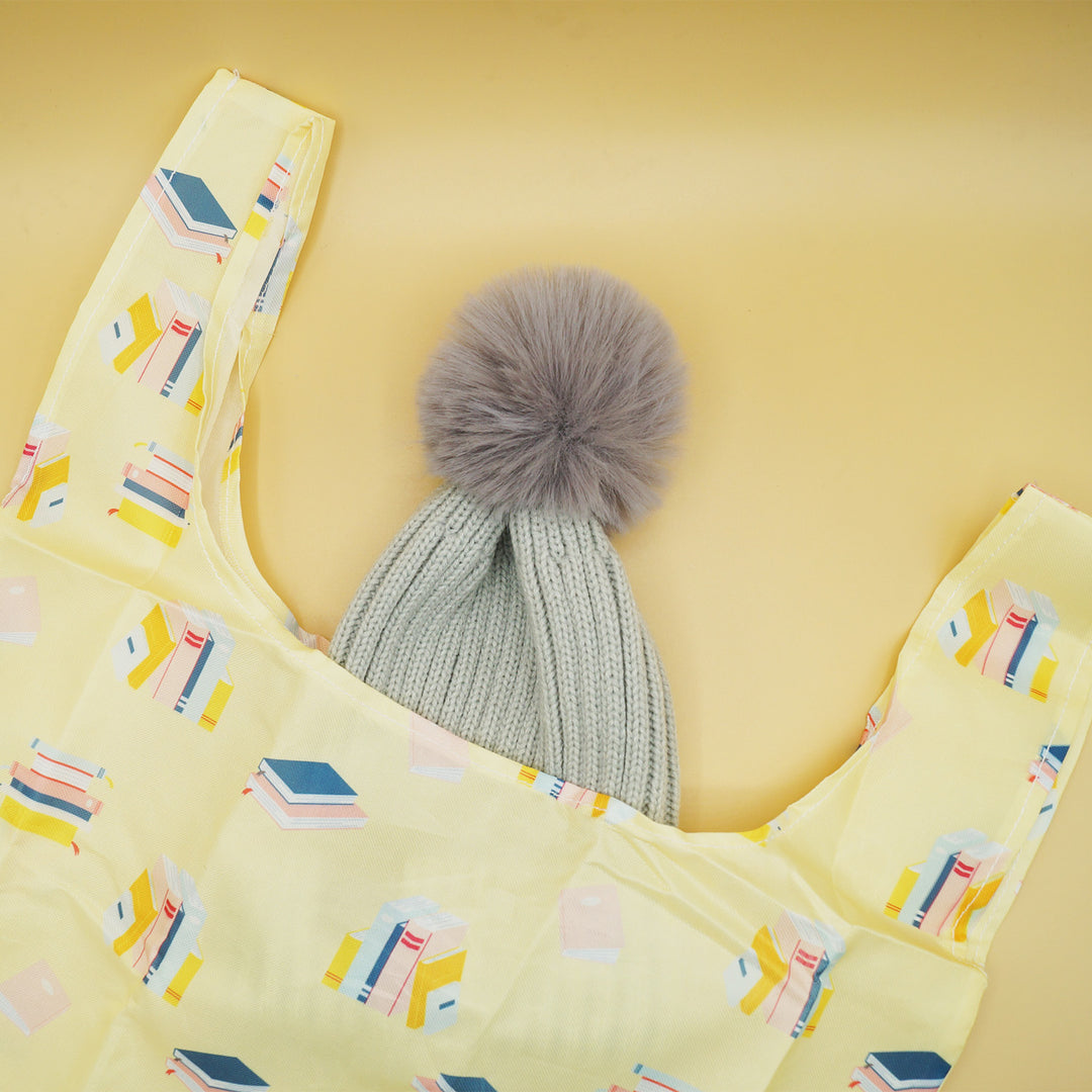 a gray pompom beanie is coming out of a yellow tote bag with books printed on it