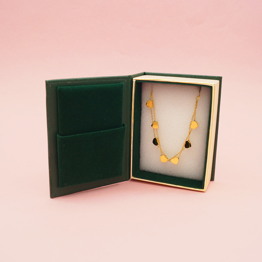 A double strand gold heart bracelet in a small book shaped box on a pink background.