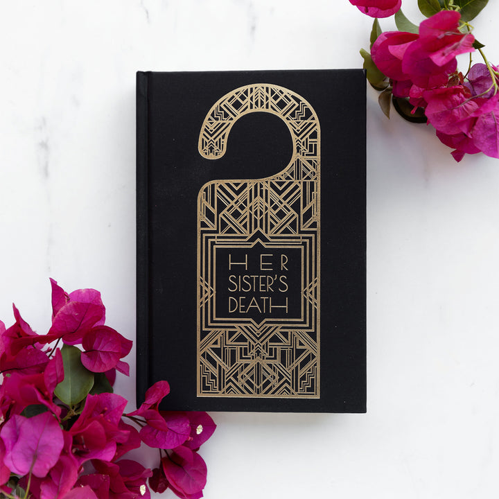 hardcover special edition of Her Sister's Death. The front is all black with a gold door hanger centered with a geometric pattern inside. Pink flowers at the bottom left and top right of the image