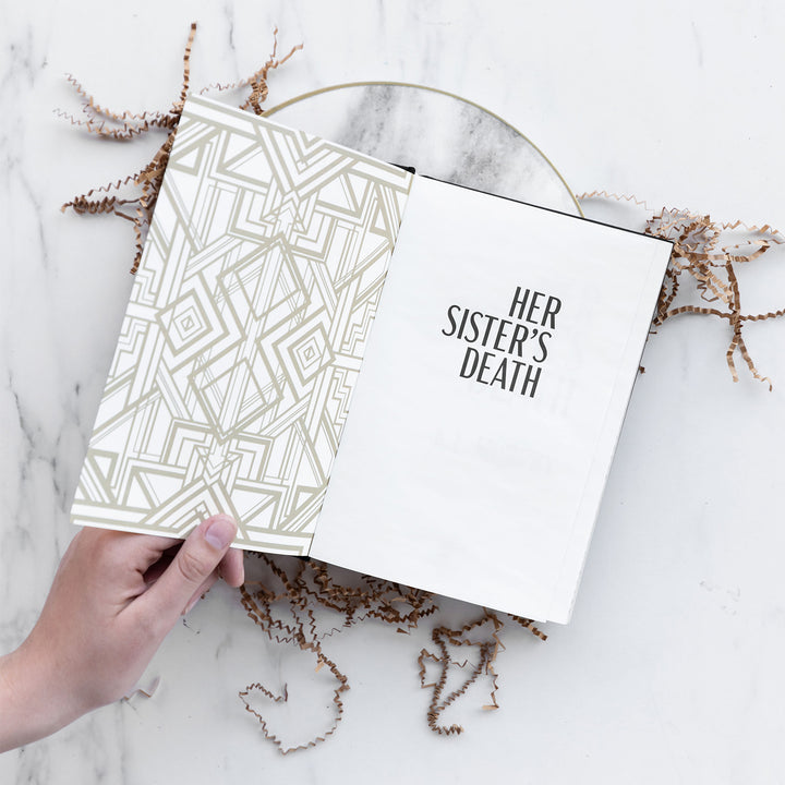 a white hand holds a hardcover special edition of Her Sister's Death open showing the title page against a marble background