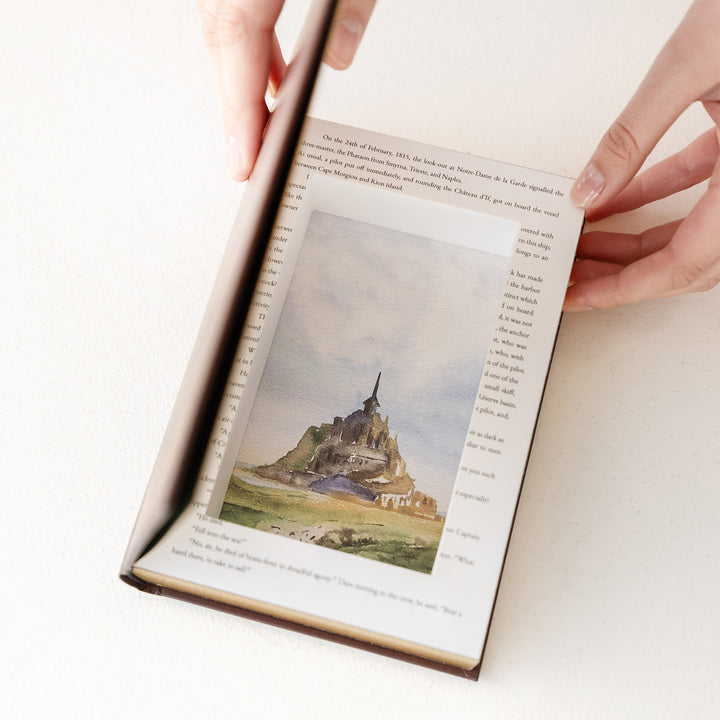 White hands hold a book open. The book has a hollow rectangle cut out of the middle and a painted scene showing a castle is shown.