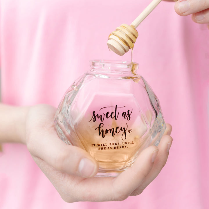 A white hand holds a clear hexagon-shaped honey jar with the words "sweet as honey. it will keep until she is ready" printed on the side and a honeycomb coming out of the top.