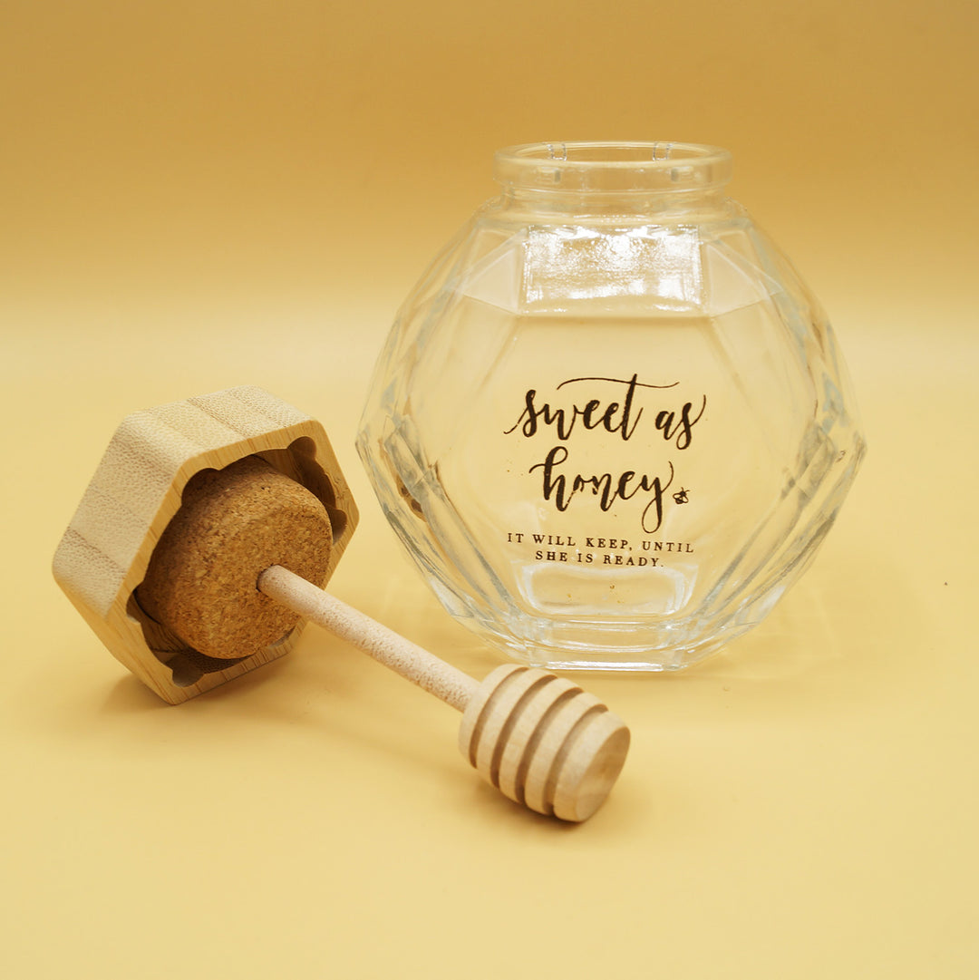 a clear hexagon-shaped honey jar with the words "sweet as honey. it will keep until she is ready" printed on the side  Wooden lid lies next to the jar and has a honeycomb attached to it