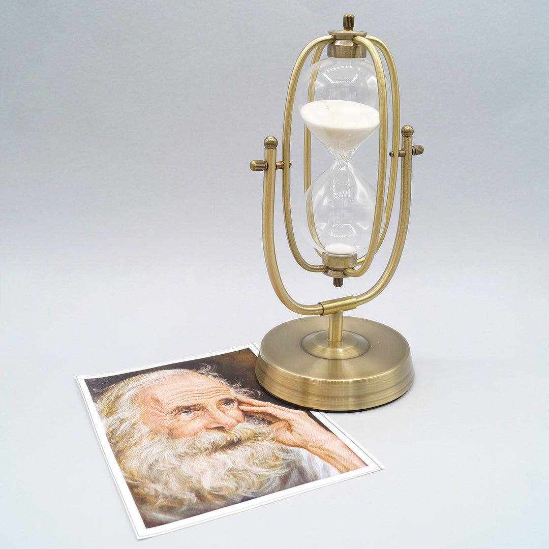 decorative, gold toned hourglass with white sand inside next to a printed image of a man with a white beard with his hand against his face.