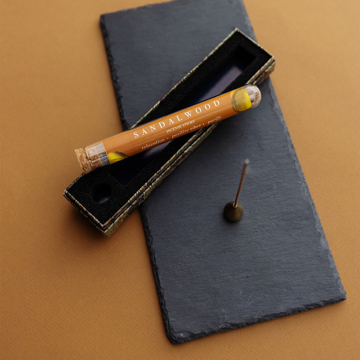 a tube of sandalwood incense sticks lays on top of an open box next to a small gold teardrop incense stick holder