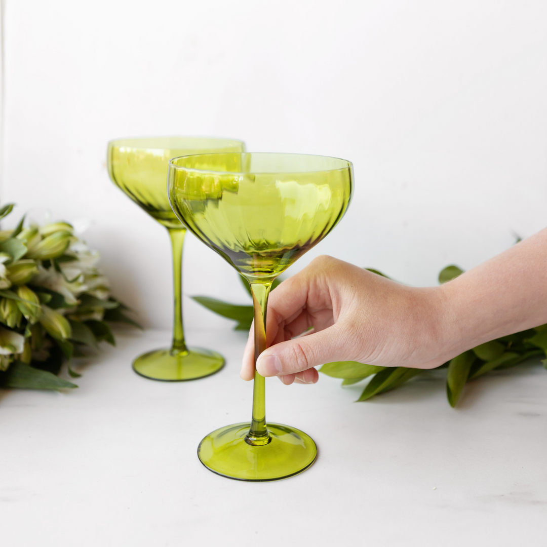 a white hand grips the base of a green coupe glass. A set of two are pictured on a white background with greenery in the background.