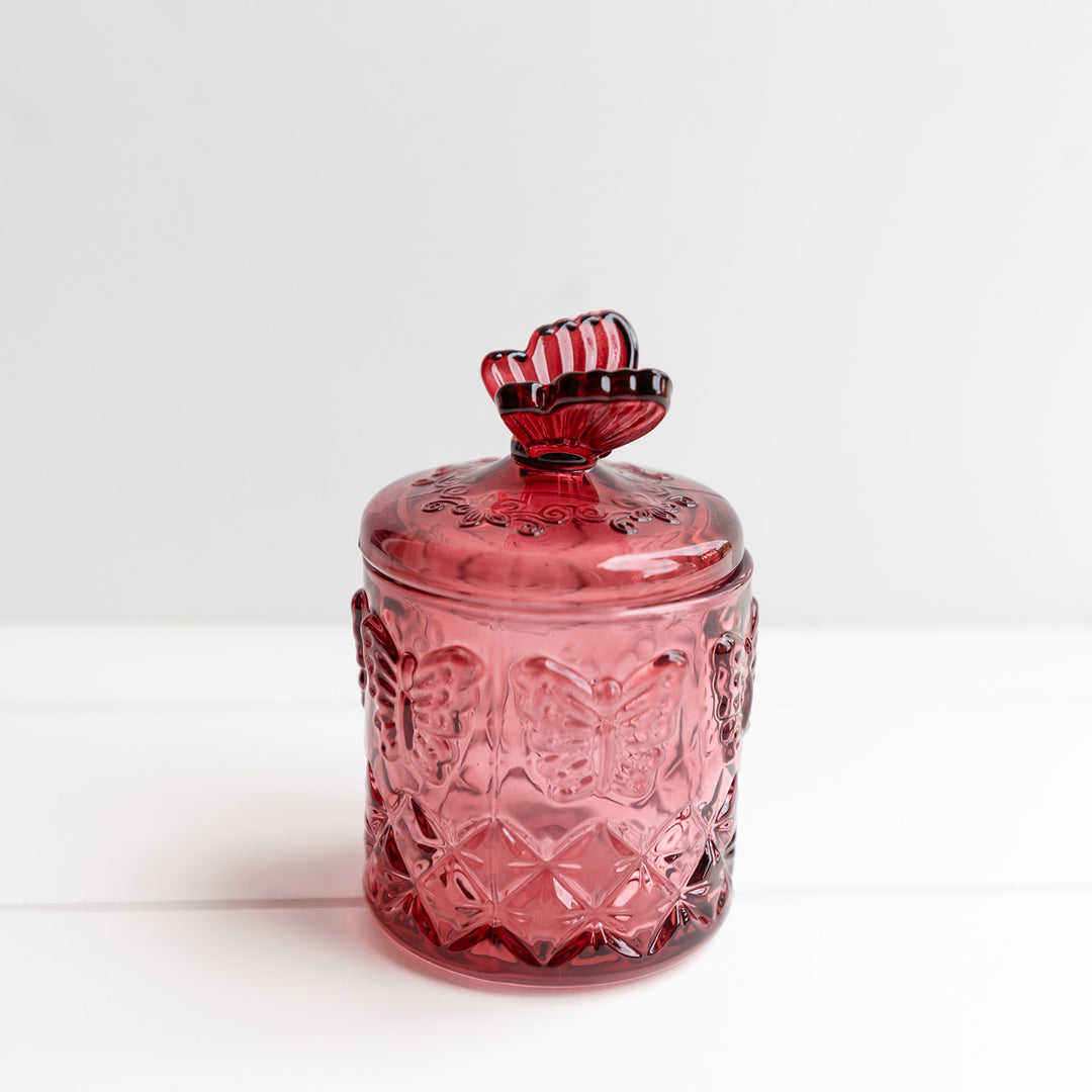 A red glass jar with a butterfly pattern on it. The lid has a red butterfly sitting on top of it.
