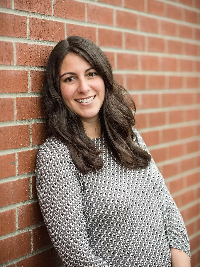 A smiling Jo Segura leans against a red brick wall, wearing a textured gray and white patterned knit top.