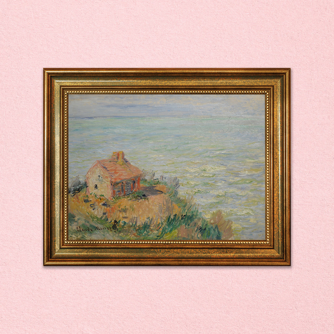 An art print on a pink background. The art print has a gold frame and shows a watercolor painting of a house on a cliff by the ocean.