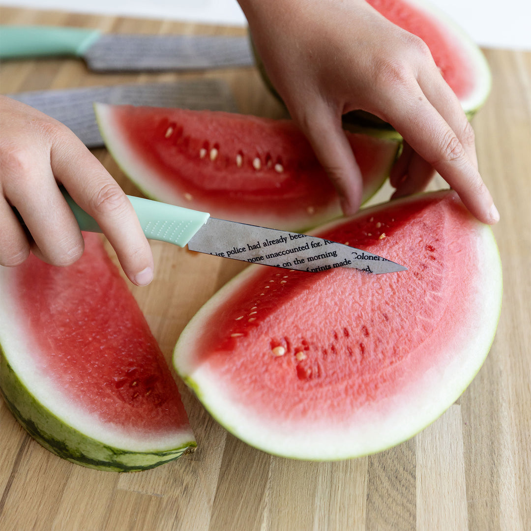 a pair of white hands hold a knife to a sliced watermelon on a wooden table. the knife has a seafoam green handle and the blade looks like pages have been pasted onto it