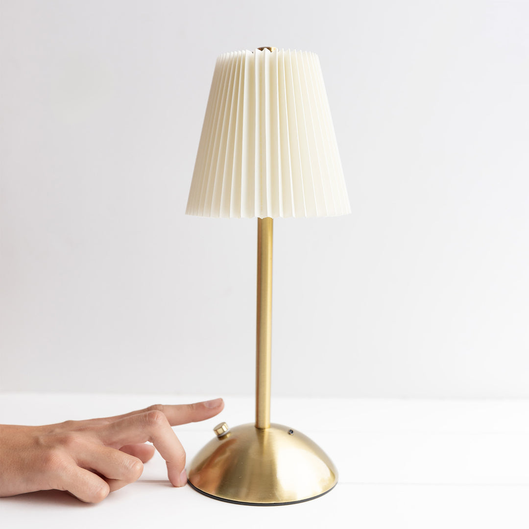 A hand reaches out to tap the base of a gold lamp. The lampshade is white and the lamp stand is gold.
