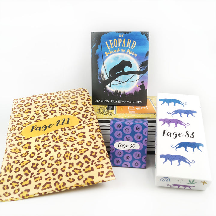 A hardcover edition of The Leopard Behind the Moon sits on a yellow box. In front are a leopard-patterned drawstring bag, a small box, and a white rectangular box. The boxes and bags all have page numbers.