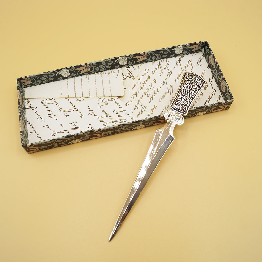 a silver letter opener with intricate design on the handle leans against the lid of a box