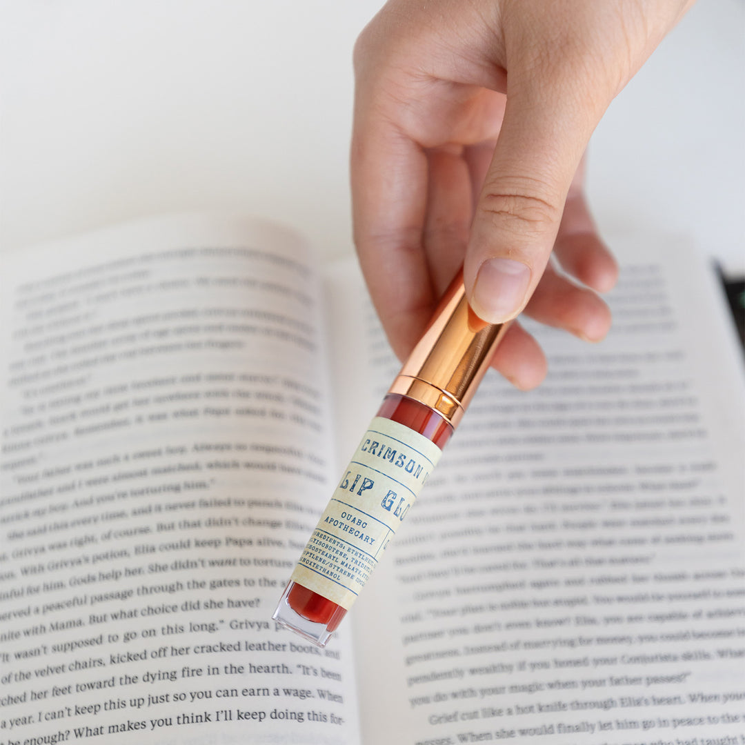 White hand holds a crimson lip gloss with a gold lid over an open book.