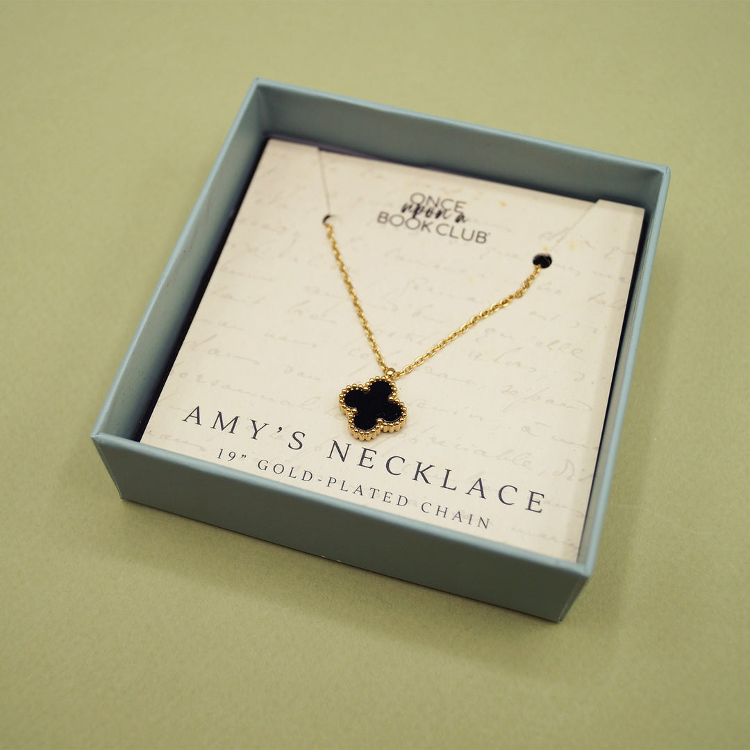 A small square open box with a gold-plated necklace. The necklace has a black cross-shaped charm on it.