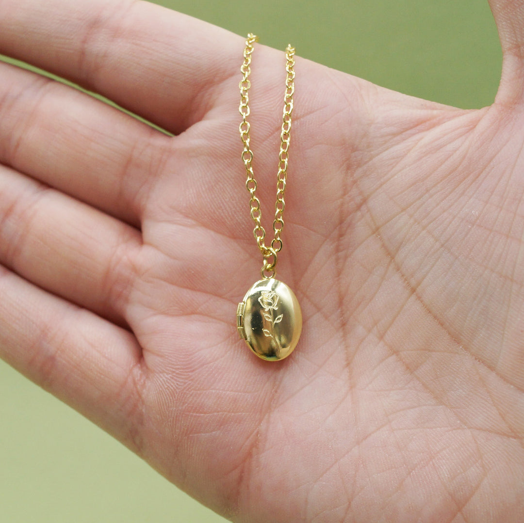 a white hand holding a gold miniature locket necklace with a rose engraved on the front