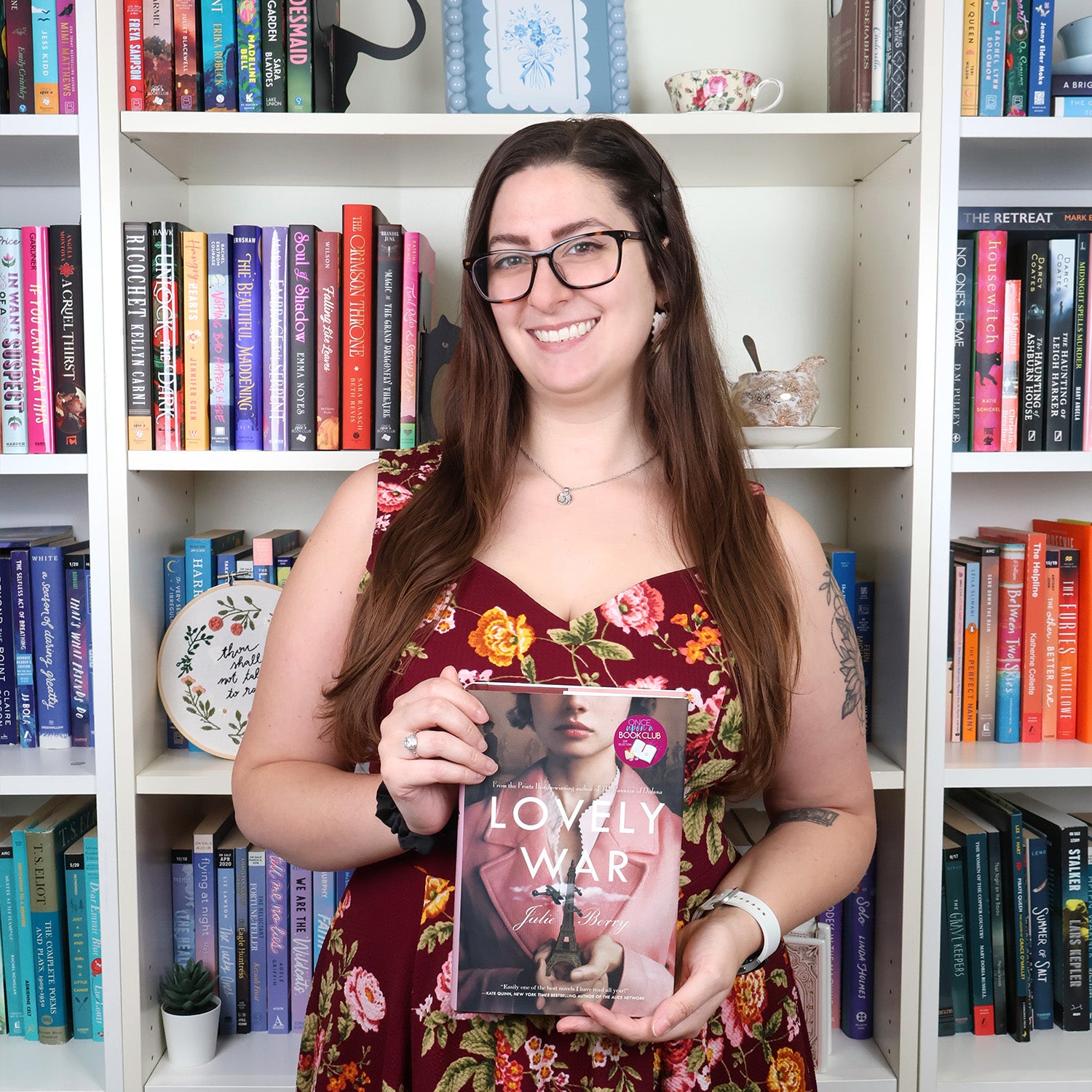 A smiling woman with long brown hair, glasses, and tattoos, wearing a floral sleeveless dress, holds a book titled 'LOVELY WAR' by Julie berry in front of white bookshelves filled with books.