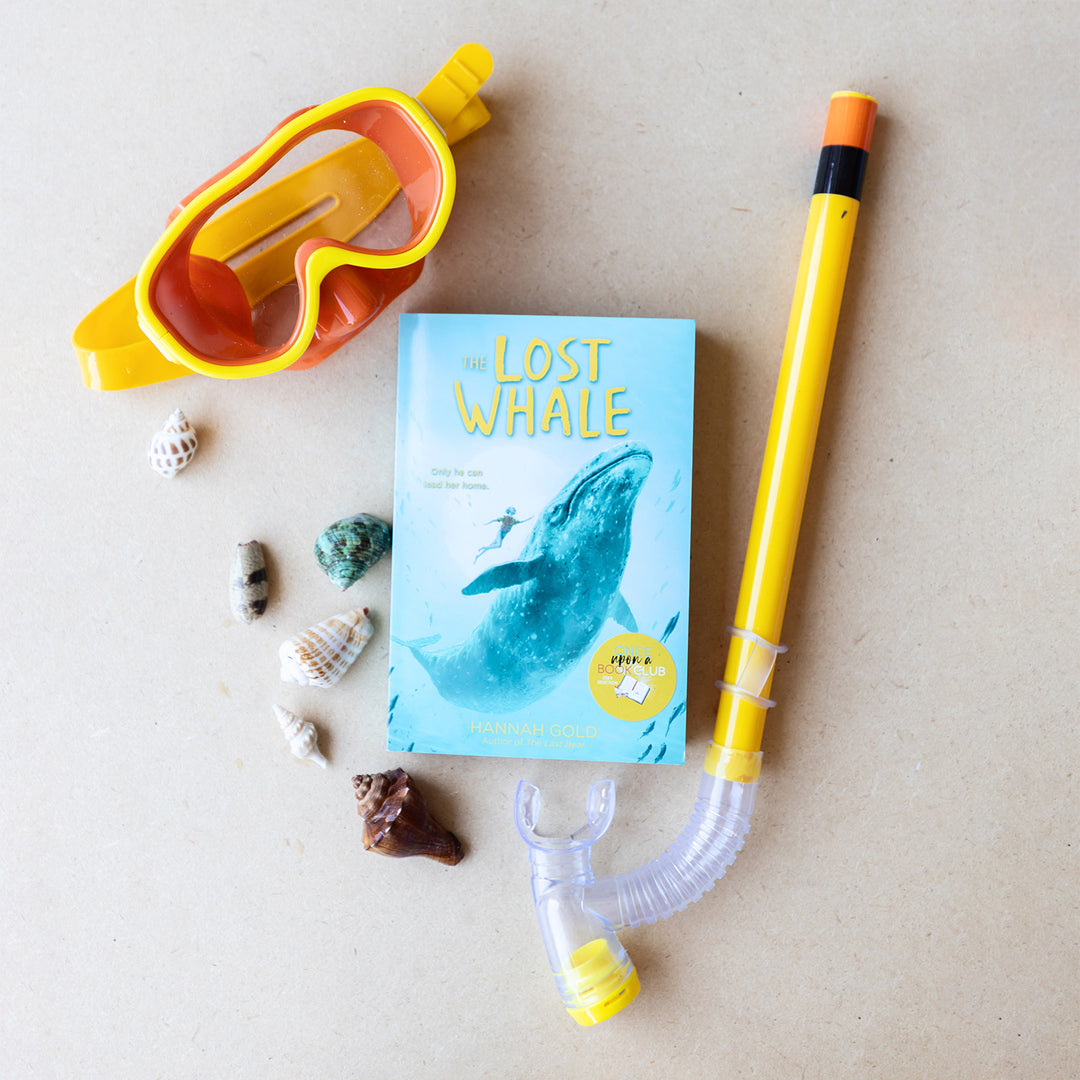 A paperback copy of The Lost Whale by Hannah Gold sits on a tan background. Surrounded by sea shells, yellow and orange diving goggles, and a snorkle.