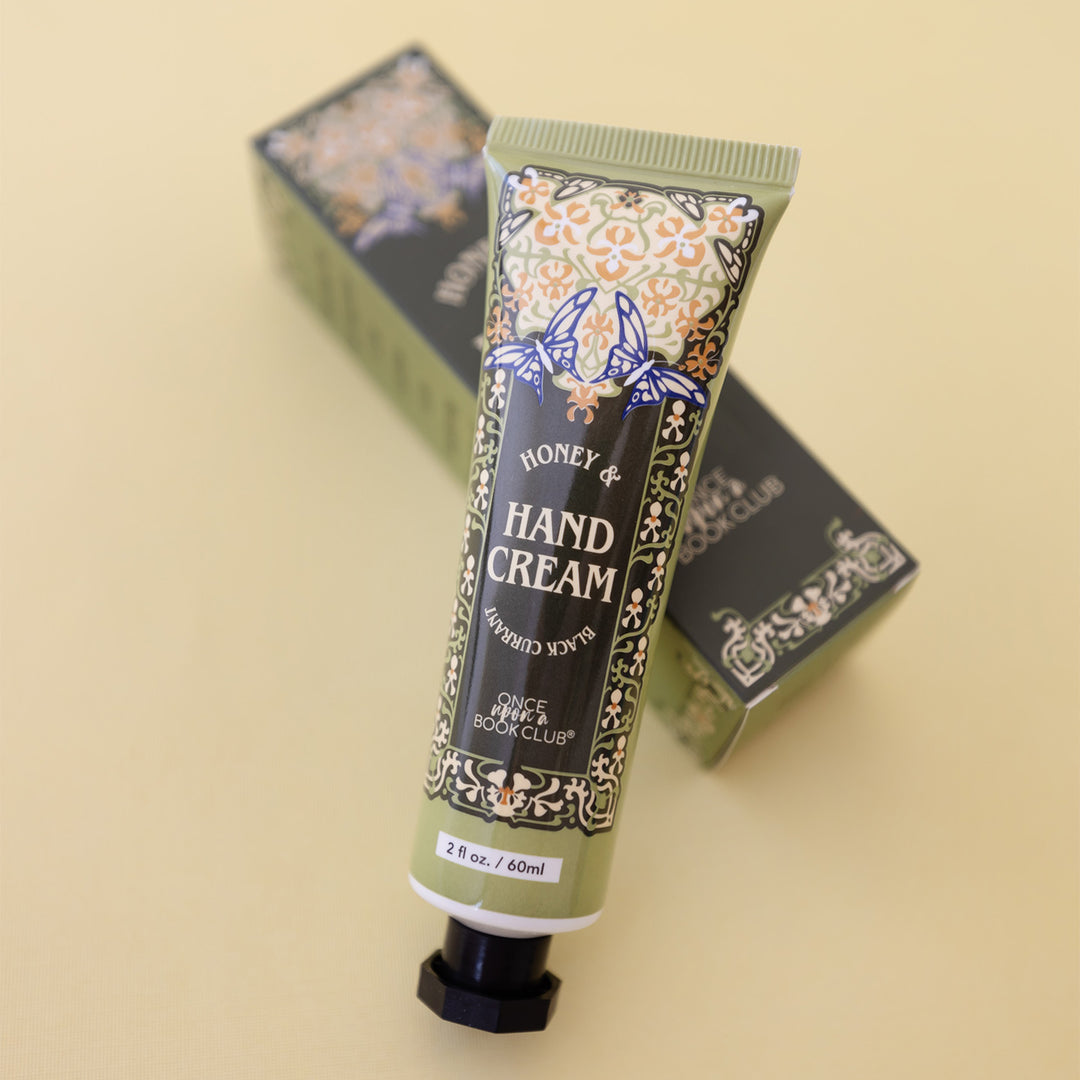 An olive green tube of Honey & Black Currant Hand Cream - Once Upon a Book Club. There is a floral pattern on the tube with two blue and white butterflies over the words. The tube lays against a matching box.