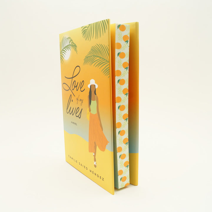 a hardcover copy of "Love of my Lives" by Yamile Saied Méndez stands slightly open with oranges printed on the page edges. On the cover, there is a beach setting and a woman faces forward in an orange skirt and green top with a white sunhat