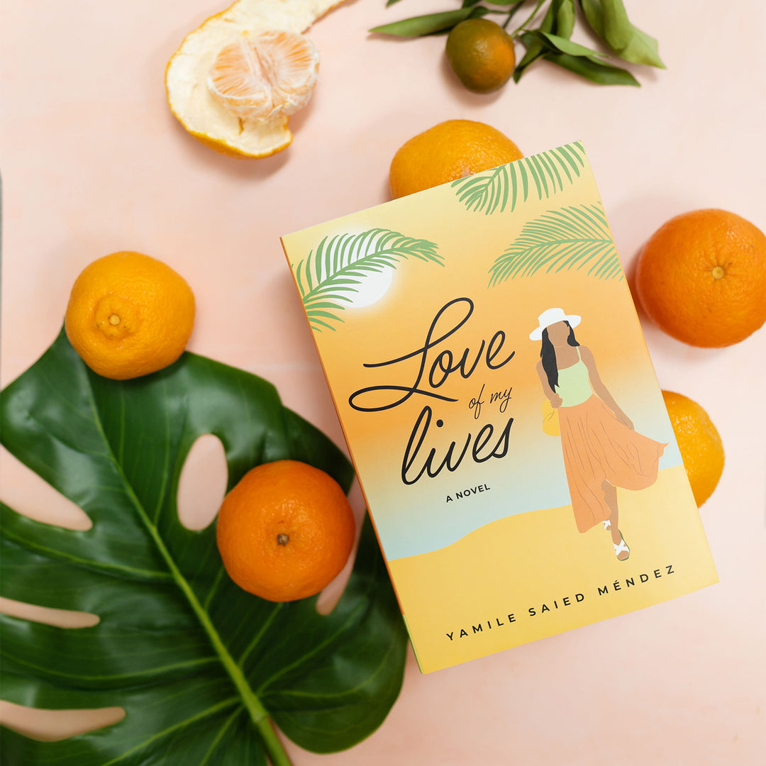 a hardcover copy of "Love of my Lives" by Yamile Saied Méndez sits on a background of oranges and palm fronds. On the cover, there is a beach setting and a woman faces forward in an orange skirt and green top with a white sunhat