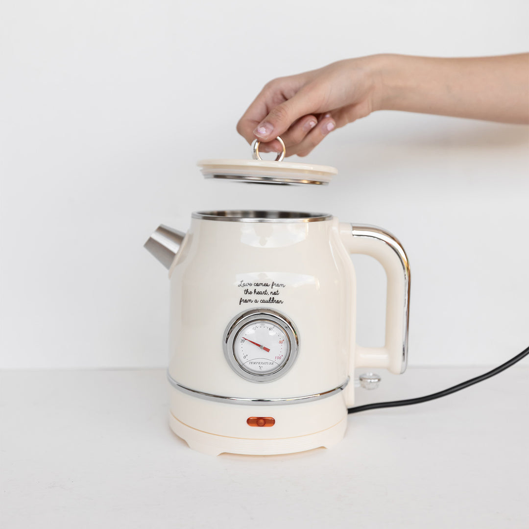 A hand holds up the lid of a cream-colored kettle. The quote on the front, above the temperature gauge, says Love comes from the heart, not from a cauldron.