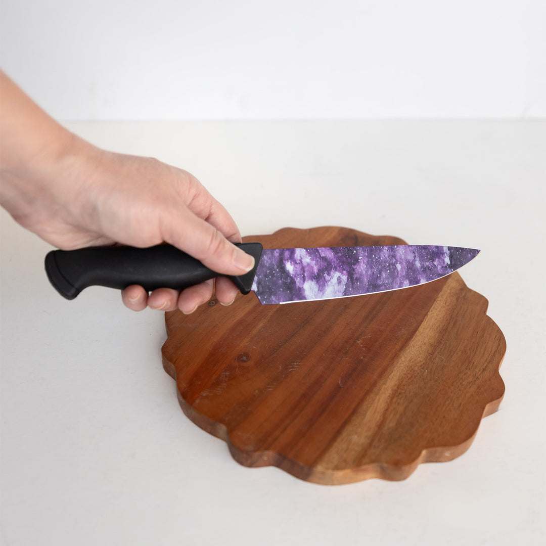 A hand holds out a kitchen knife with a black handle and galaxy-print on the blade above a wooden circular board.