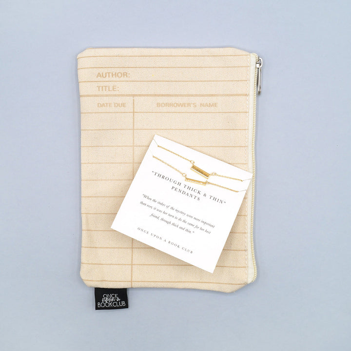 a beige library card patterned makeup bag with a set of two gold-plated bar necklaces on top, labeled "Through Thick & Thin Pendant"