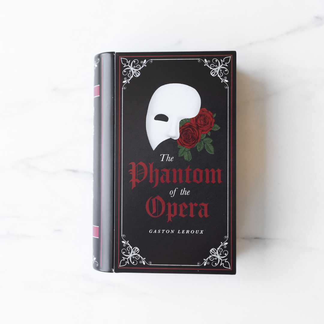 A book shaped tin sits on a white marble background. The book tin is black and designed to look like a copy of The Phantom of the Opera by Gaston Leroux.